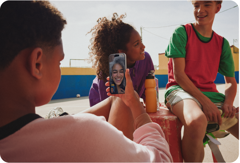 Um grupo de crianças sentadas ao ar livre. Uma criança está em uma ligação do FaceTime em um iPhone.