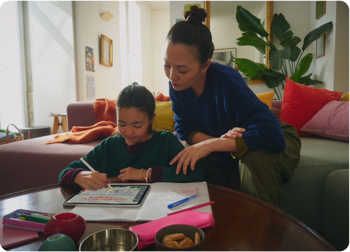 Vienas iš tėvų ir vaikas sėdi prie stalo, vaikas piešia „iPad“ naudodamas „Apple&nbsp;Pencil“.
